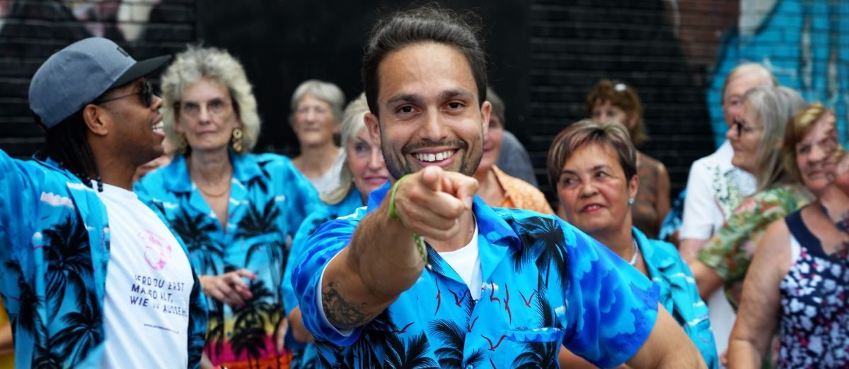 Ein junger Mann in blauem, gemustertem Hemd zeigt mit dem Finger. Im Hintergrund Senior*innen.