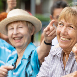 Seniorin zeigt lächelnd in die Kamera. Hinter ihr eine Gruppe weiterer Senior*innen, tanzend.