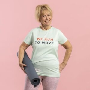 Eine sportliche Seniorin hält eine zusammengerollte Yogamatte unter dem Arm und lächelt in die Kamera.