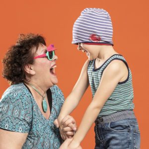 Eine ältere Frau und ein Kind lachen sich herzlich an und halten sich an den Händen. Beide tragen bunte Kleidung – das Kind hat die Mütze spielerisch über die Augen gezogen. Der orange Hintergrund verstärkt die fröhliche, lebendige Stimmung.