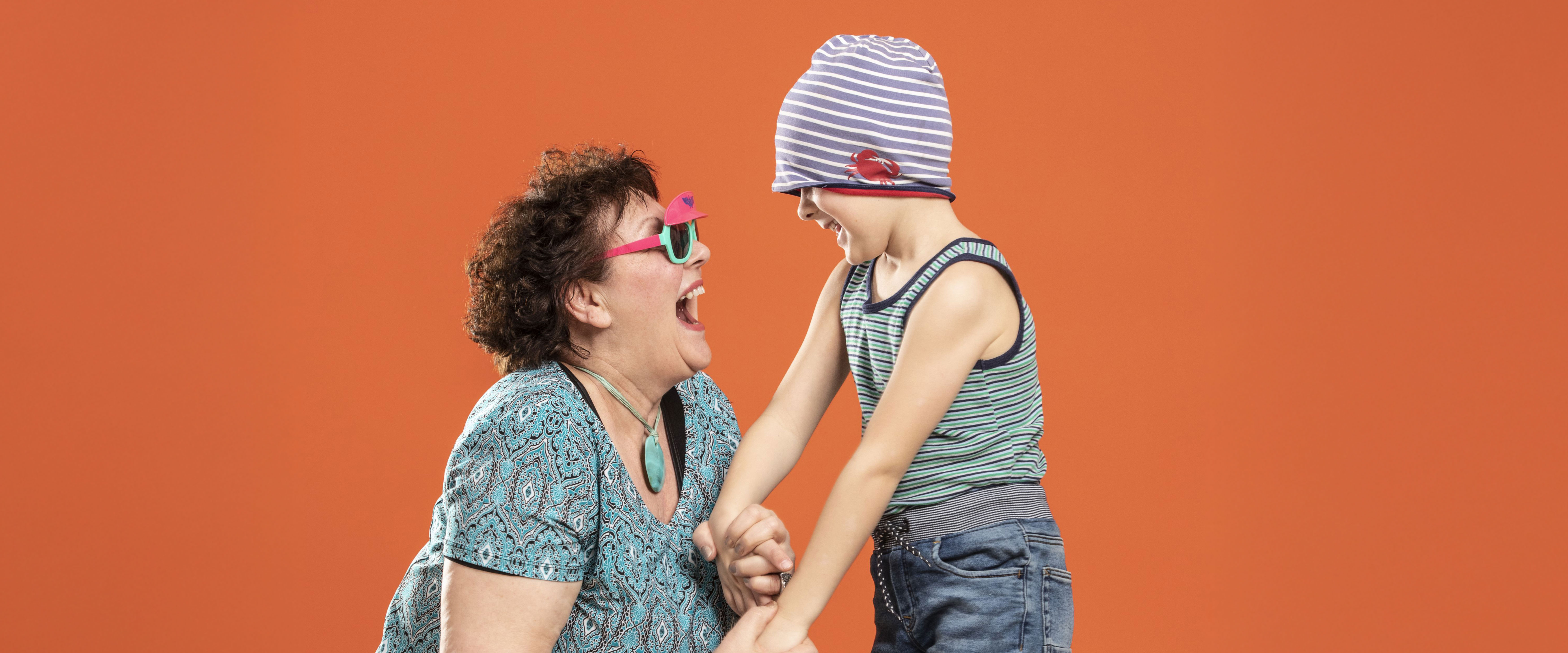 Eine ältere Frau und ein Kind lachen sich herzlich an und halten sich an den Händen. Beide tragen bunte Kleidung – das Kind hat die Mütze spielerisch über die Augen gezogen. Der orange Hintergrund verstärkt die fröhliche, lebendige Stimmung.