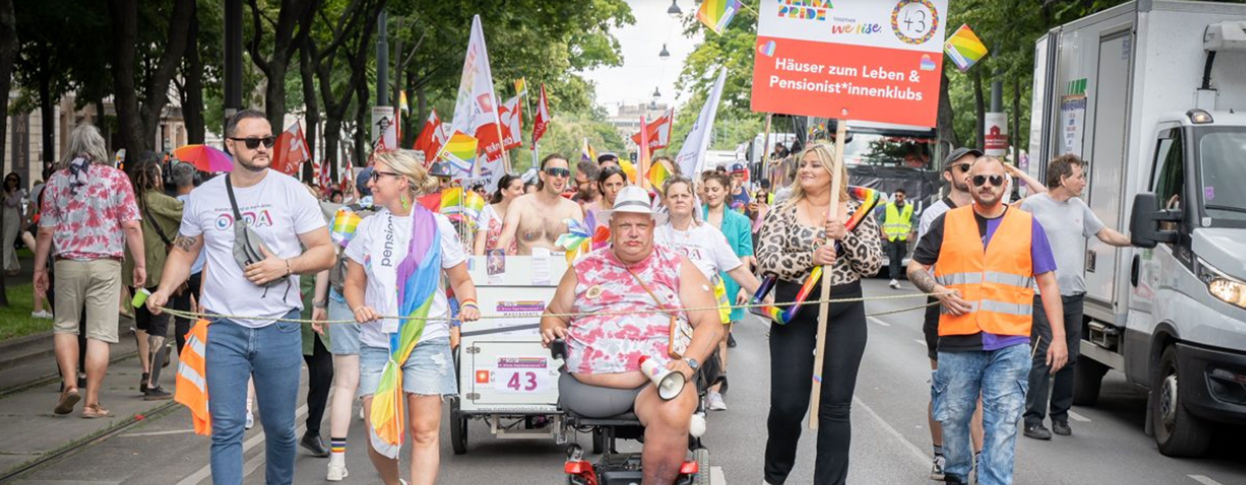 Das Bild zeigt eine Gruppe von Menschen, die bei einer Parade mitlaufen. Es handelt sich um eine bunte und lebhafte Szene im Freien, vermutlich im Rahmen einer Pride-Veranstaltung, wie die Regenbogenfahnen und -accessoires andeuten.