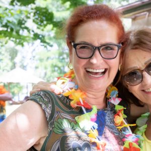 zwei Seniorinnen stehen im Freien, lächeln in die Kamera und tragen Blumenketten