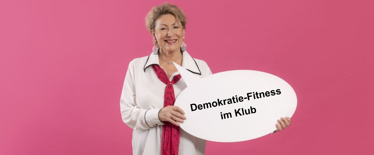 Seniorin hält eine Papp-Sprechblase mit der Aufschrift "Demokratie-Fitness im Klub" in die Kamera.