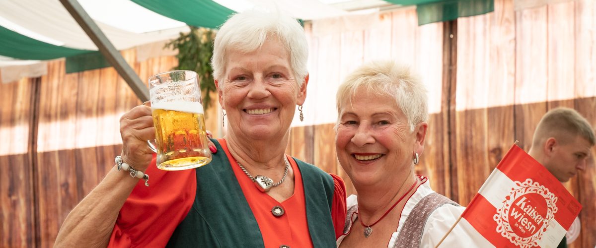 Zwei Seniorinnen in Tracht im Festzelt halten einen Bierkrug und ein Werbefähnchen lächelnd in die Kamera.