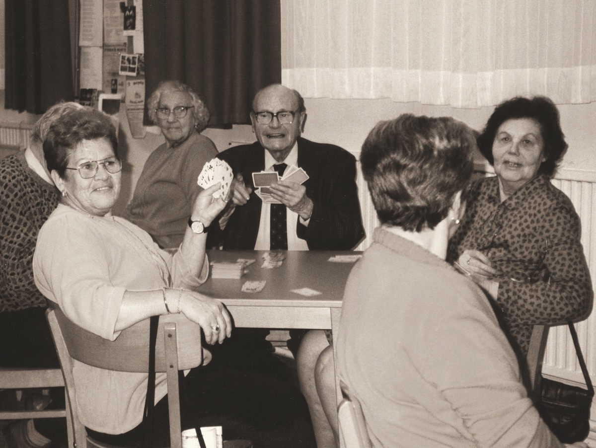 Historisches schwarzweiß-Foto eines Pensionistenklubs aus 1960, mehrere Senior*innen sitzen an beinem Tisch und spielen Karten.