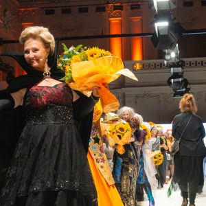 Seniorin mit prachtvollem Designer-Outfit und Blumenstrauß in der Hand schreitet lächelnd über den Laufsteg der Vienna Fashion Week. Weitere Models und begeistertes Publikum im Hintergrund.