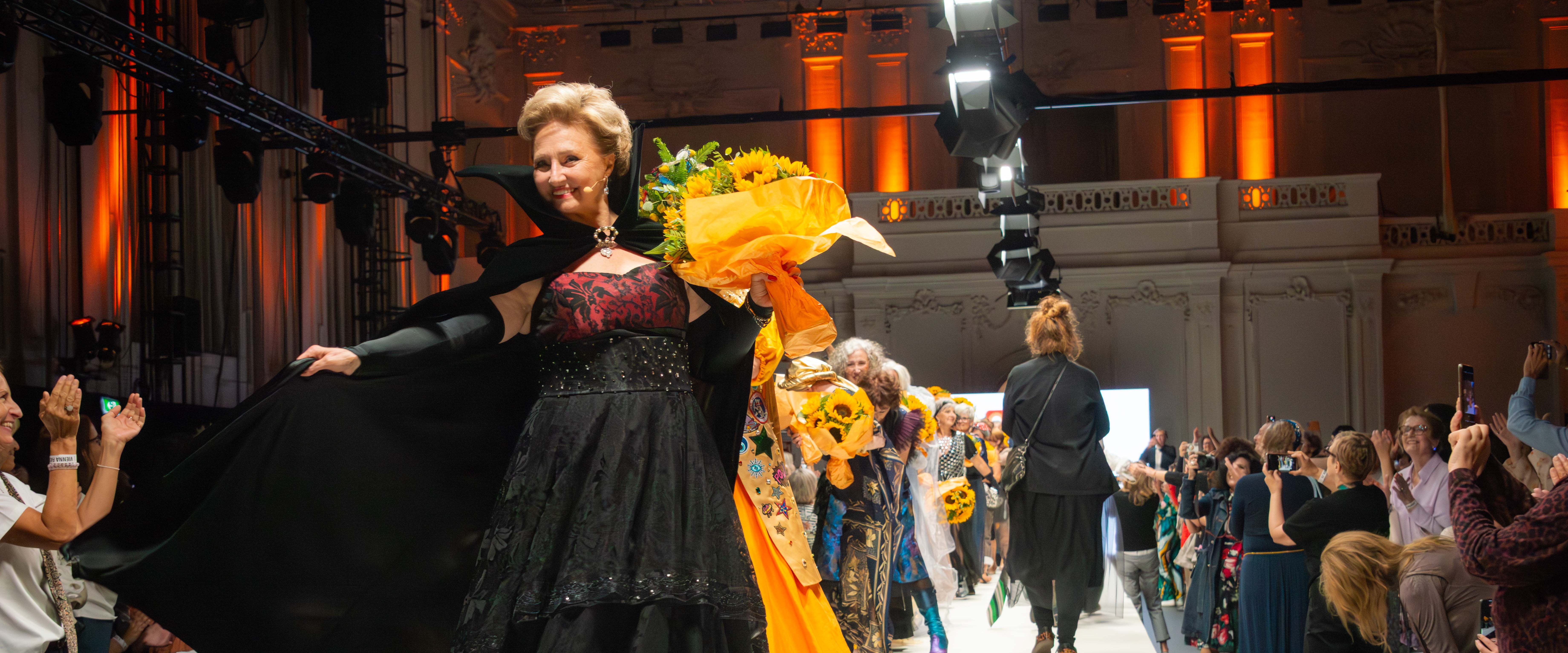 Seniorin mit prachtvollem Designer-Outfit und Blumenstrauß in der Hand schreitet lächelnd über den Laufsteg der Vienna Fashion Week. Weitere Models und begeistertes Publikum im Hintergrund.