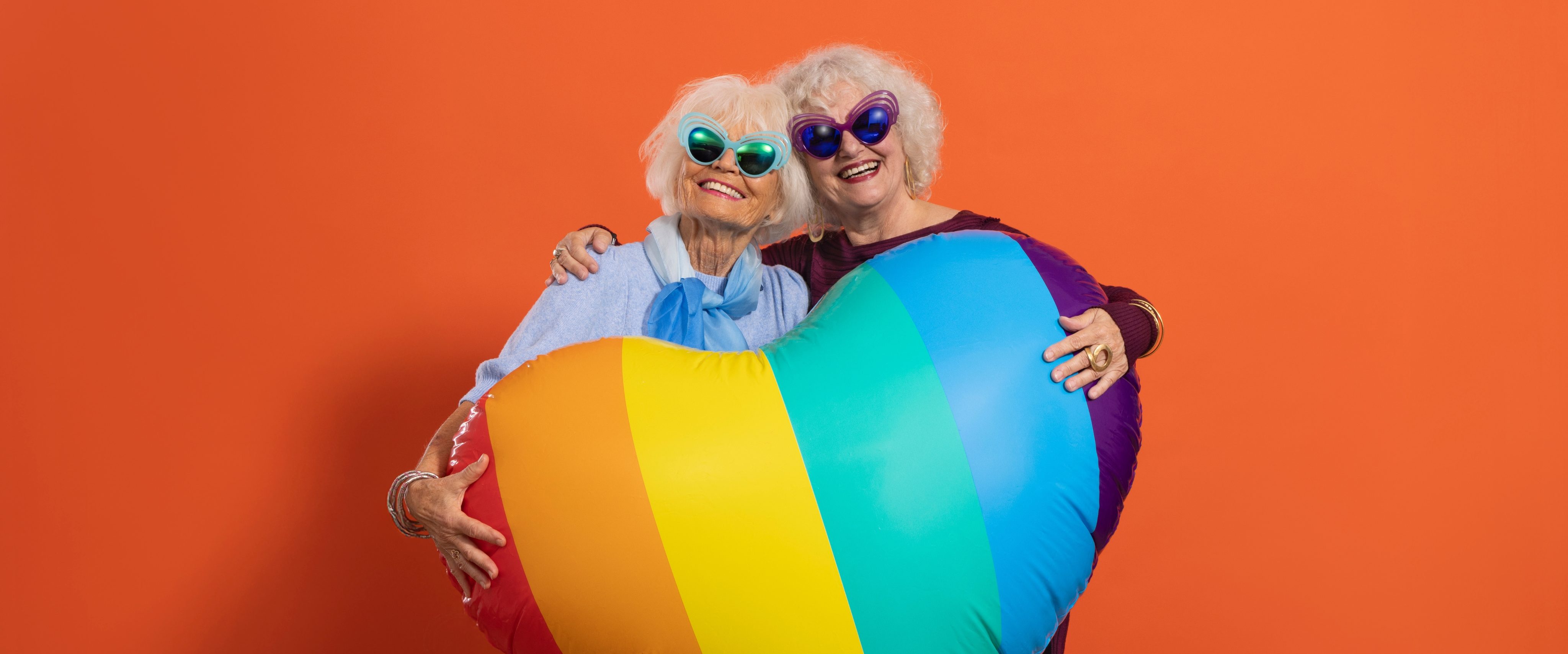 Zwei Senior*innen mit bunten Sonnenbrillen halten gemeinsam ein großes Regenbogen-Herz.
