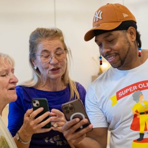 Zwei Seniorinnen und ein jüngerer Mann mit Kappe und lustigem T-Shirt it Handys.