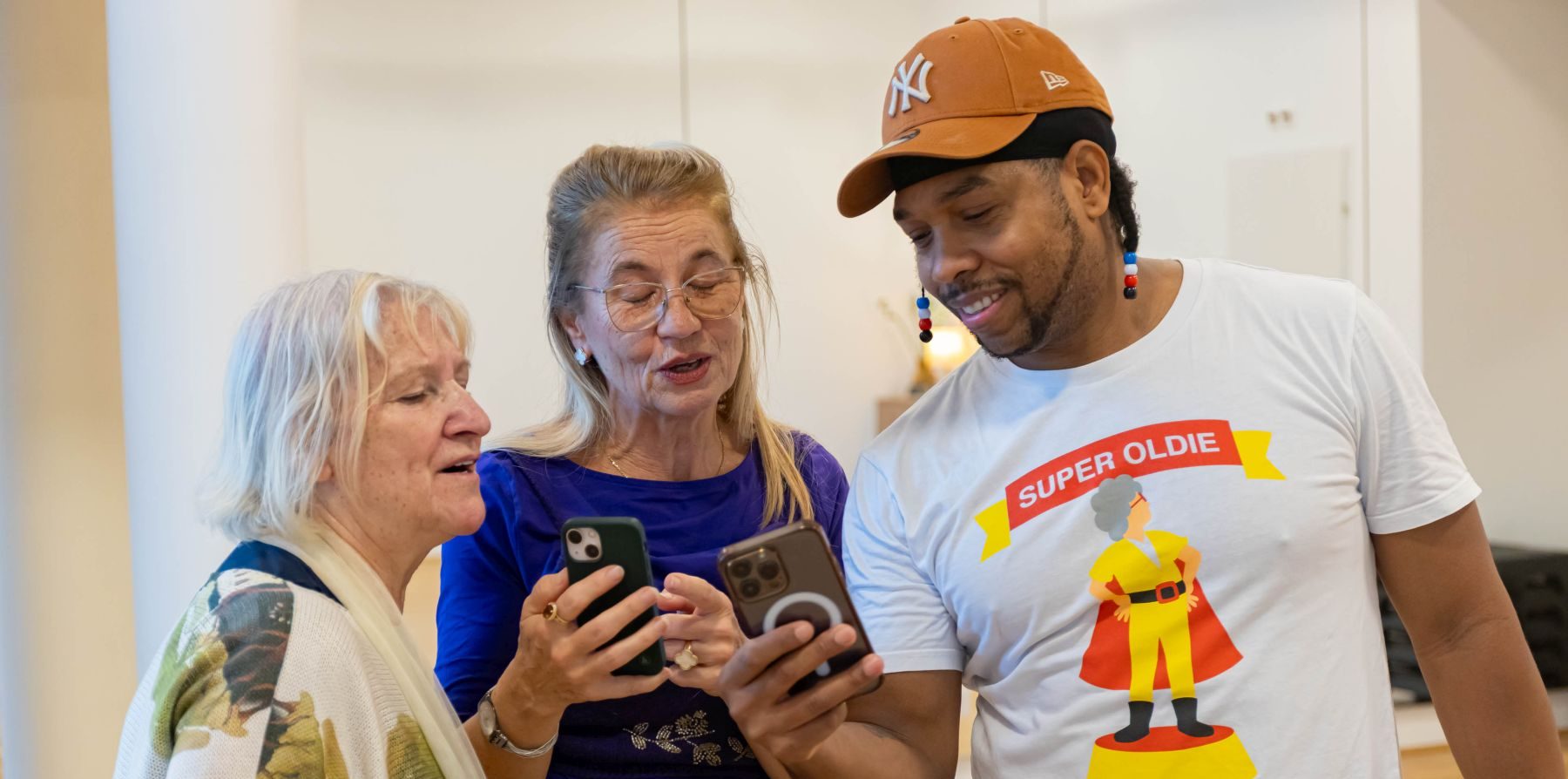 Zwei Seniorinnen und ein jüngerer Mann mit Kappe und lustigem T-Shirt it Handys.