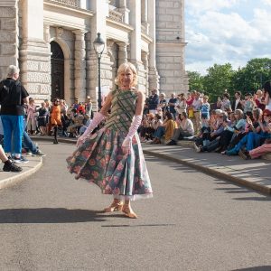 Eine ältere Frau präsentiert ein weites, langes Kleid vor dem Burgtheater in Wien im Rahmen der Klima Biennale. Rechts und links sitzen applaudierende Zuschauer*innen.