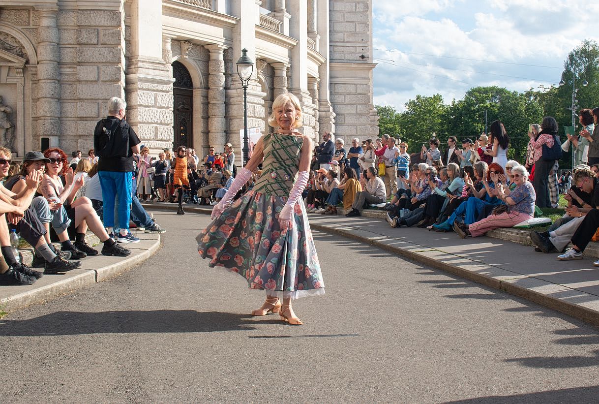Eine ältere Frau präsentiert ein weites, langes Kleid vor dem Burgtheater in Wien im Rahmen der Klima Biennale. Rechts und links sitzen applaudierende Zuschauer*innen.