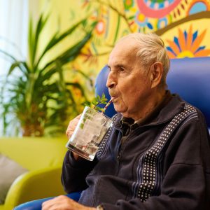 Ein älterer Herr sitzt auf einem blauen Feauteil und trinkt aus einem Glas mit Strohhalm ein Getränk mit Eiswürfeln.