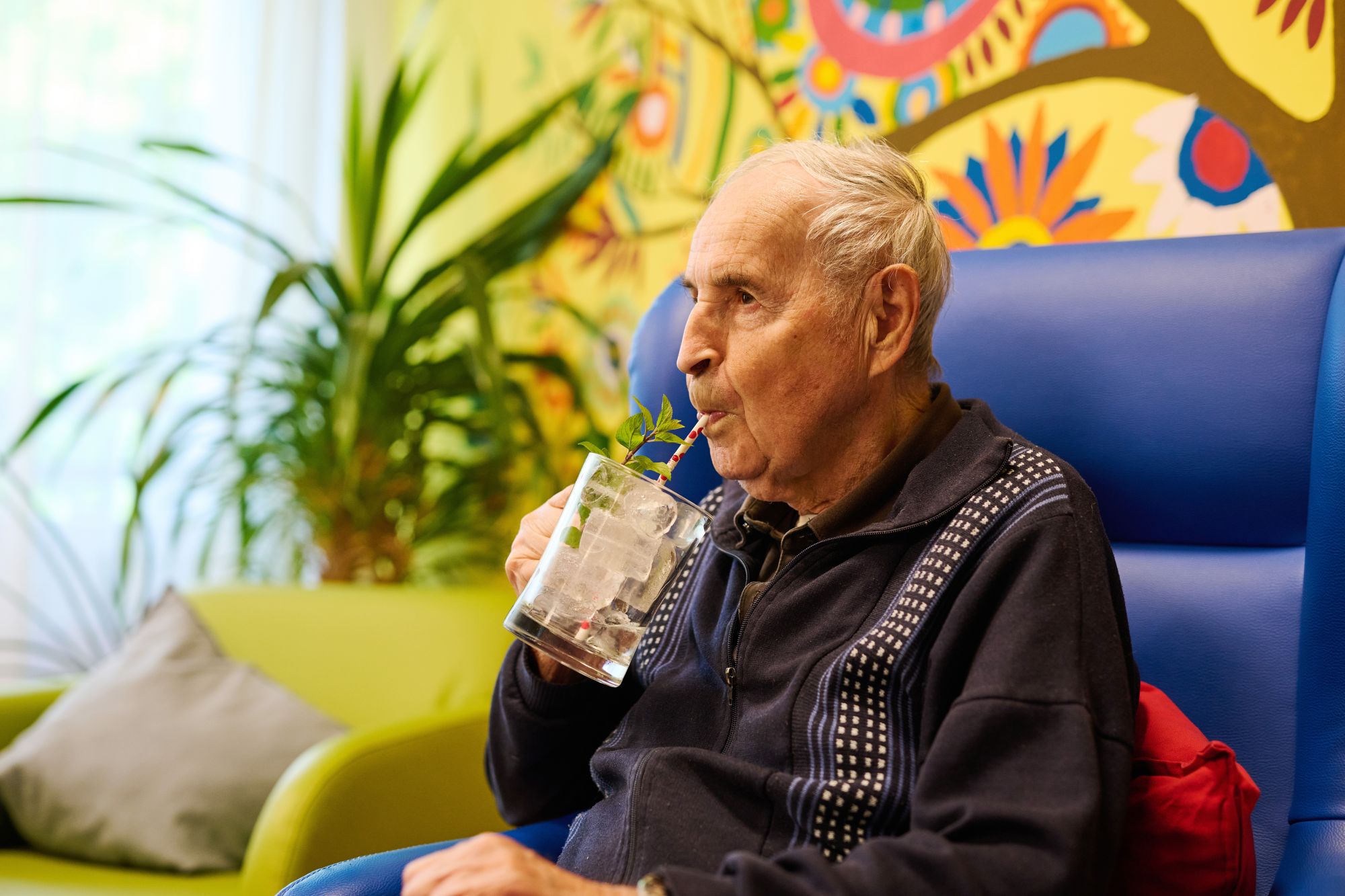 Ein älterer Herr sitzt auf einem blauen Feauteil und trinkt aus einem Glas mit Strohhalm ein Getränk mit Eiswürfeln.