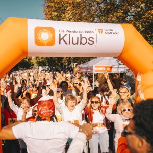 Zahlreiche Teilnehmer*innen am Nordic Walking Day 2026 stehen dicht gedrängt, heben die Arme, bei einem orangefarbenen Bogen mit Logo und Aufschrift "Pensionist*innenklubs für die Stadt Wien"