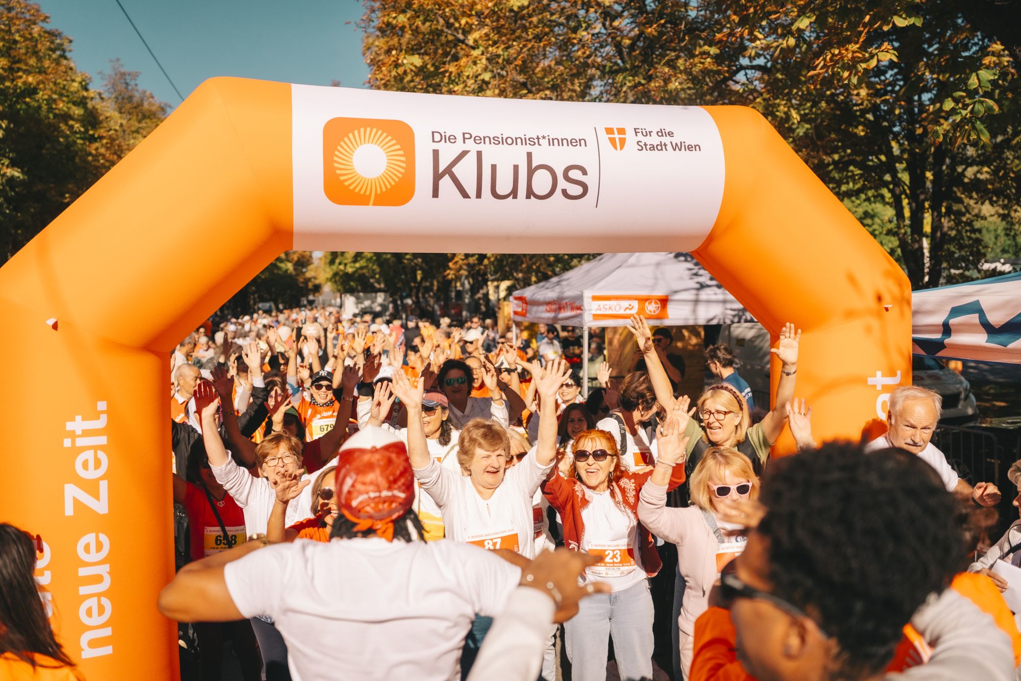 Zahlreiche Teilnehmer*innen am Nordic Walking Day 2026 stehen dicht gedrängt, heben die Arme, bei einem orangefarbenen Bogen mit Logo und Aufschrift "Pensionist*innenklubs für die Stadt Wien"