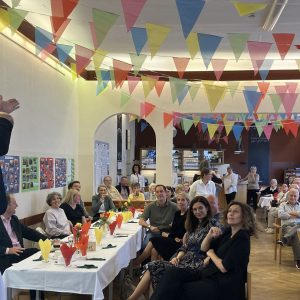 Mehrere Menschen sitzen an Tischen mit weißen Tischtüchern, ein Mann steht auf einem Sessel und gebärdet. An der Decke hängen bunte Wimpel.