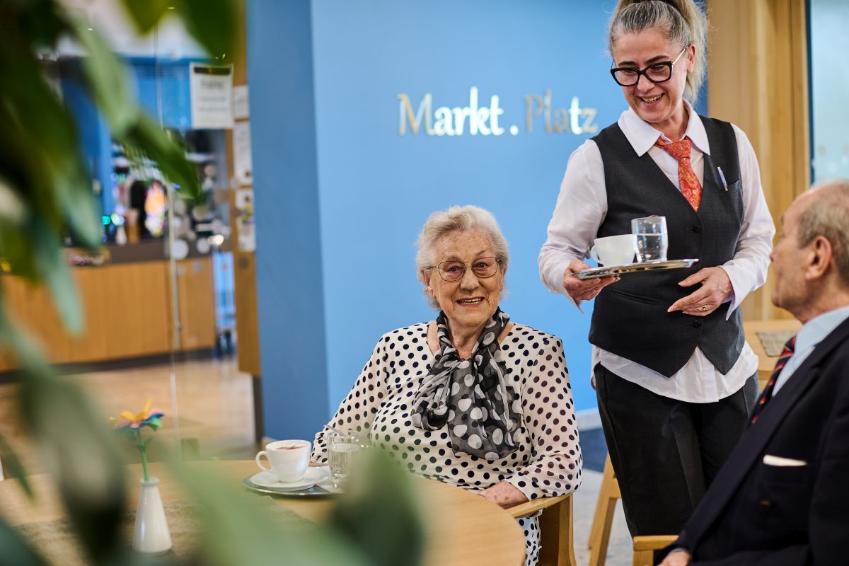 Weibliche Servicekraft serviert Kaffee, Seniorin und Senior sitzen am Tisch am "Markt.Platz".