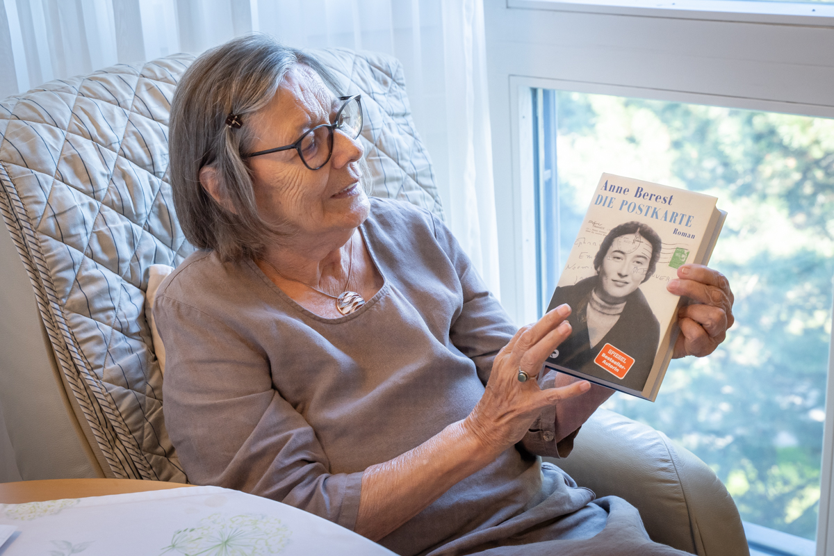 Eine ältere Frau mit grauen Haaren, Pagenkopf und Brille hält das Buch mit dem Titel "Die Postkarte" in der Hand.