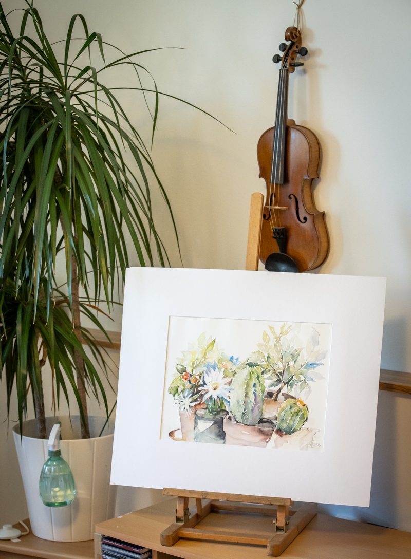 Ein Gemälde mit Blumen und Kakteen steht auf einer Kommode, daneben steht eine Palme, dahinter hängt eine Geige an der Wand.