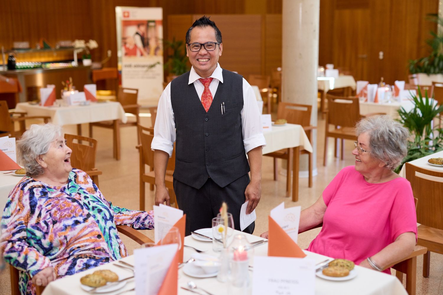 Ein Kellner und zwei Seniorinnen an einem Tisch im Demenz-Restaurant.