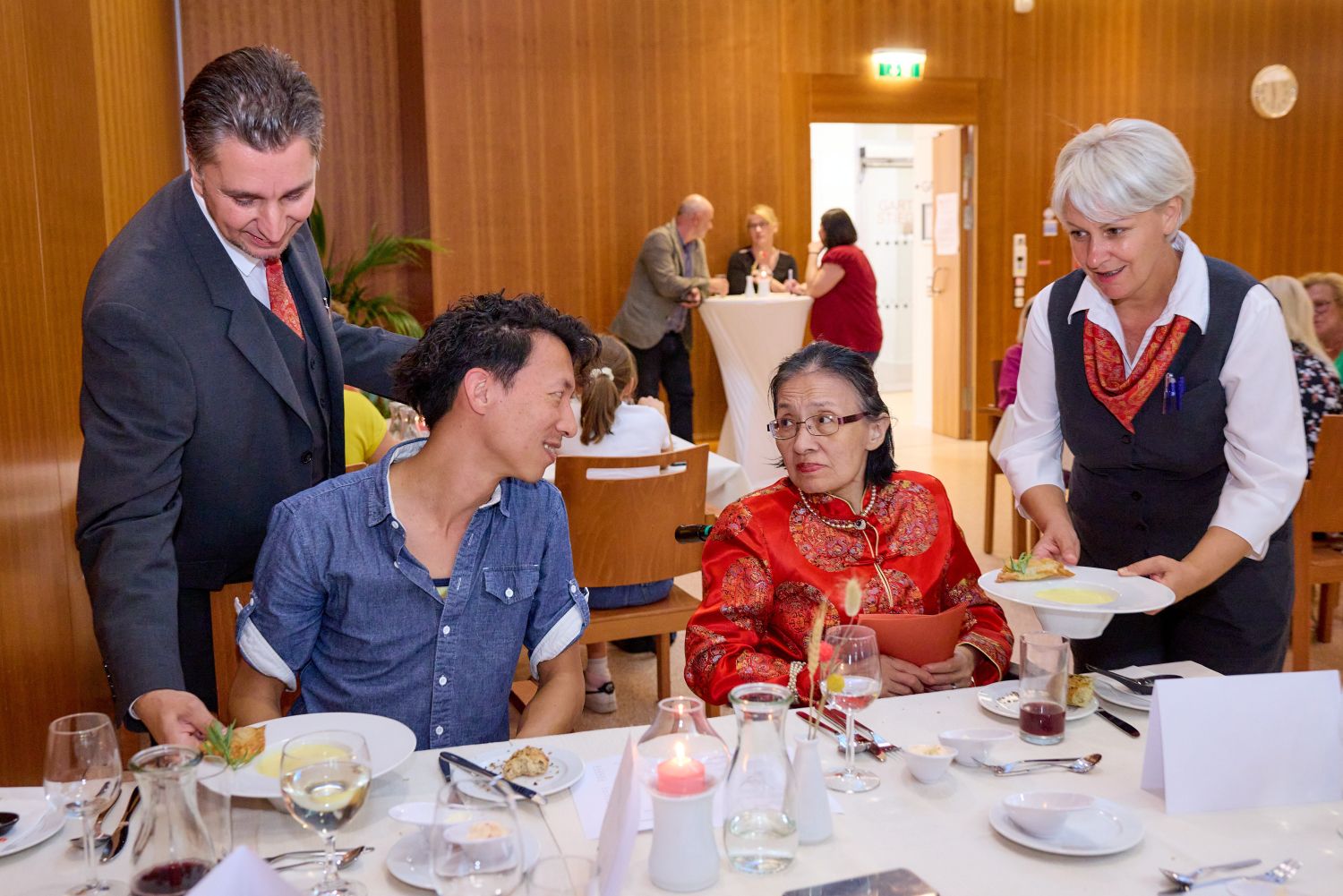 Ein junger Mann und ein ältere Frau in rotem Kleid sitzen am Tisch, während sie von zwei Servicekräften bedient werden.