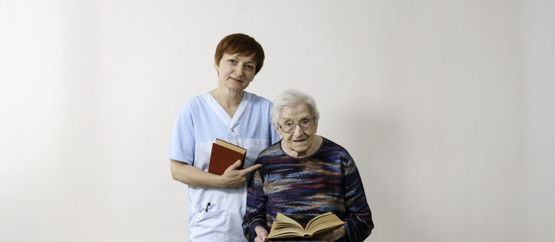 Eine Pflegerin und eine Seniorin mit einem Stapel an Büchern auf einem Rollator