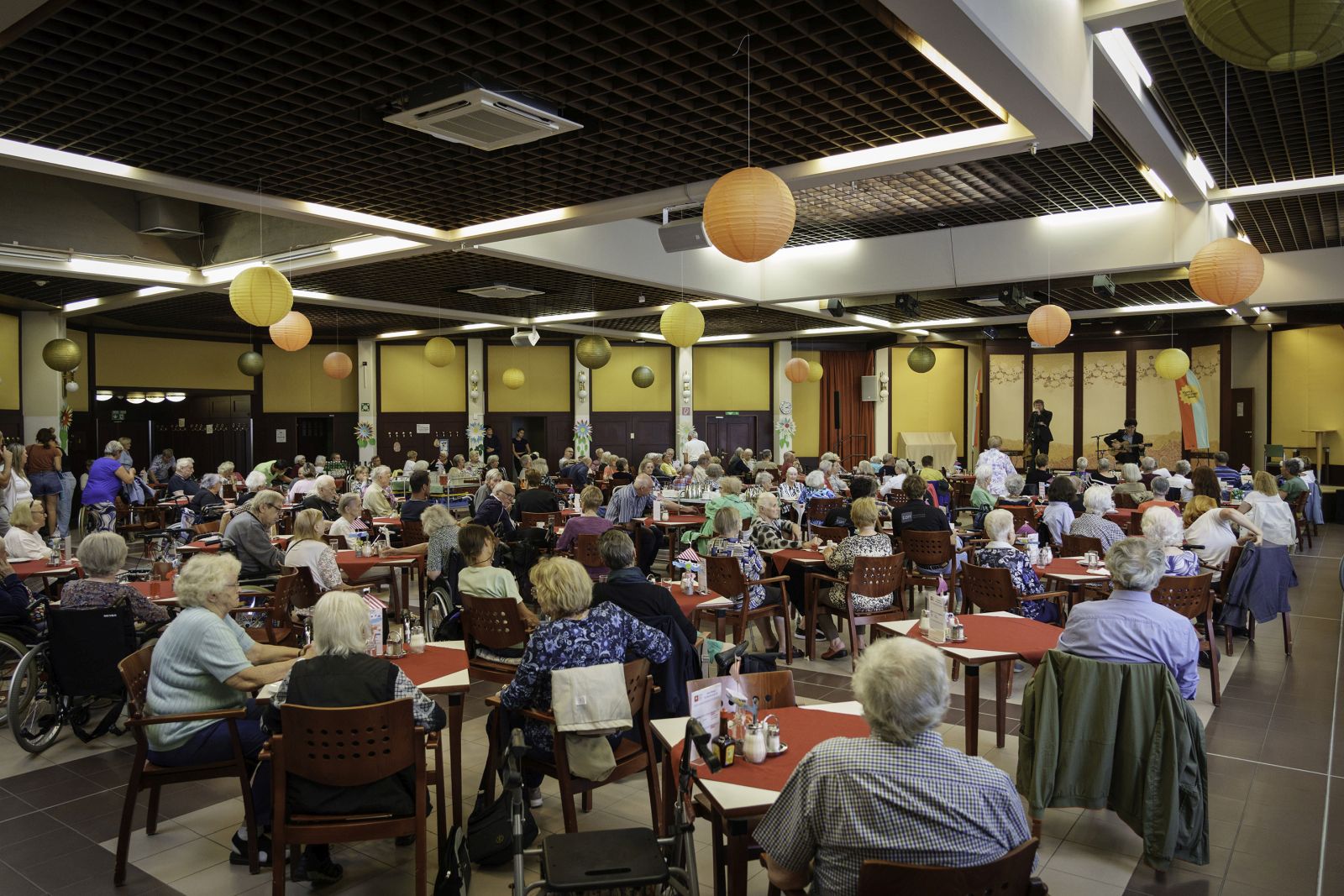 Senior*innen an Tischen im gut gefüllten GastHaus während eines Konzertes in einem Pensionisten-Wohnhaus der Häuser zum Leben.