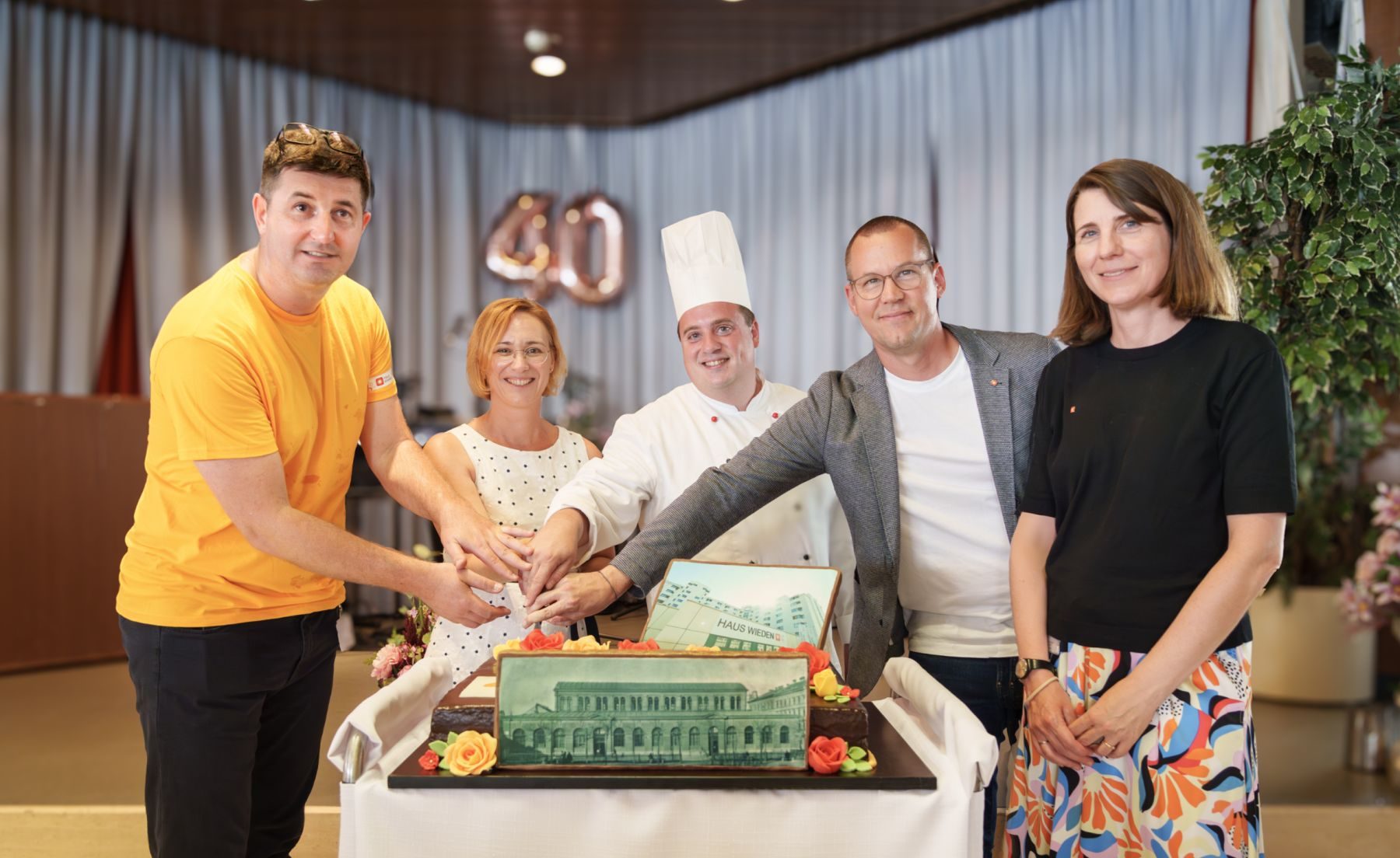 Armin Cehic, Direktor Haus Wieden, Lea Halbwidl, Bezirksvorsteherin Wieden, ein Koch, Simon Bluma, stellvertretender Geschäftsführer Häuser zum Leben und Antonia Schwarz, Regionalleiterin Häuser, Häuser zum Leben, schneiden eine Torte an.