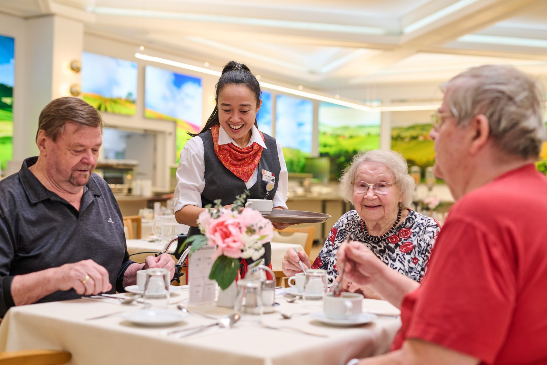 Eine Servicekraft bedient zwei Senioren und eine Seniorin an einem Tisch im GastHaus im Pensiionisten-Wohnhaus Am Mühlengrund in Wien-Liesing.
