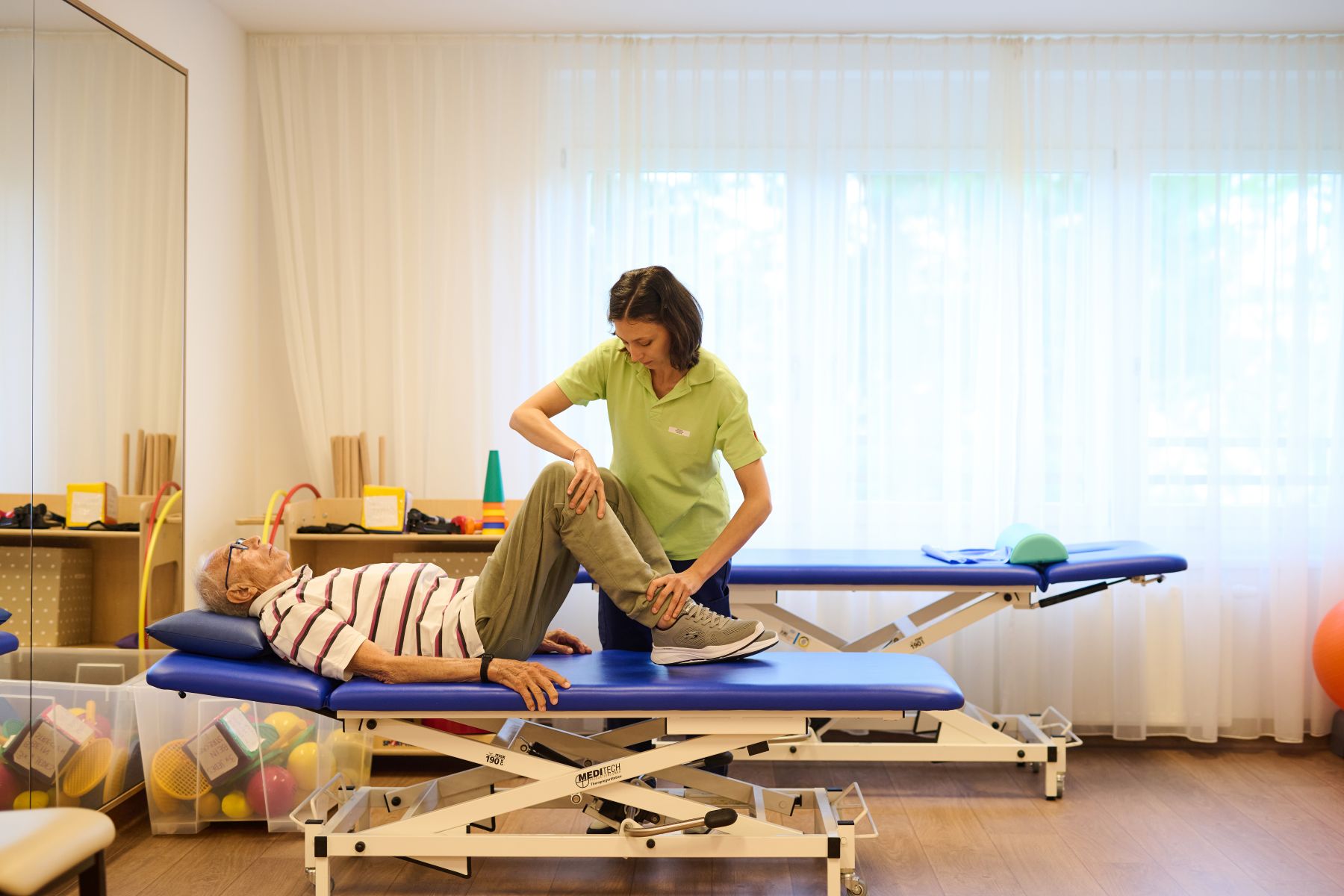 Eine Physiotherapeutin neben einem älteren Herrn, der mit abgewinkeltem Knie auf einer Liege liegt.