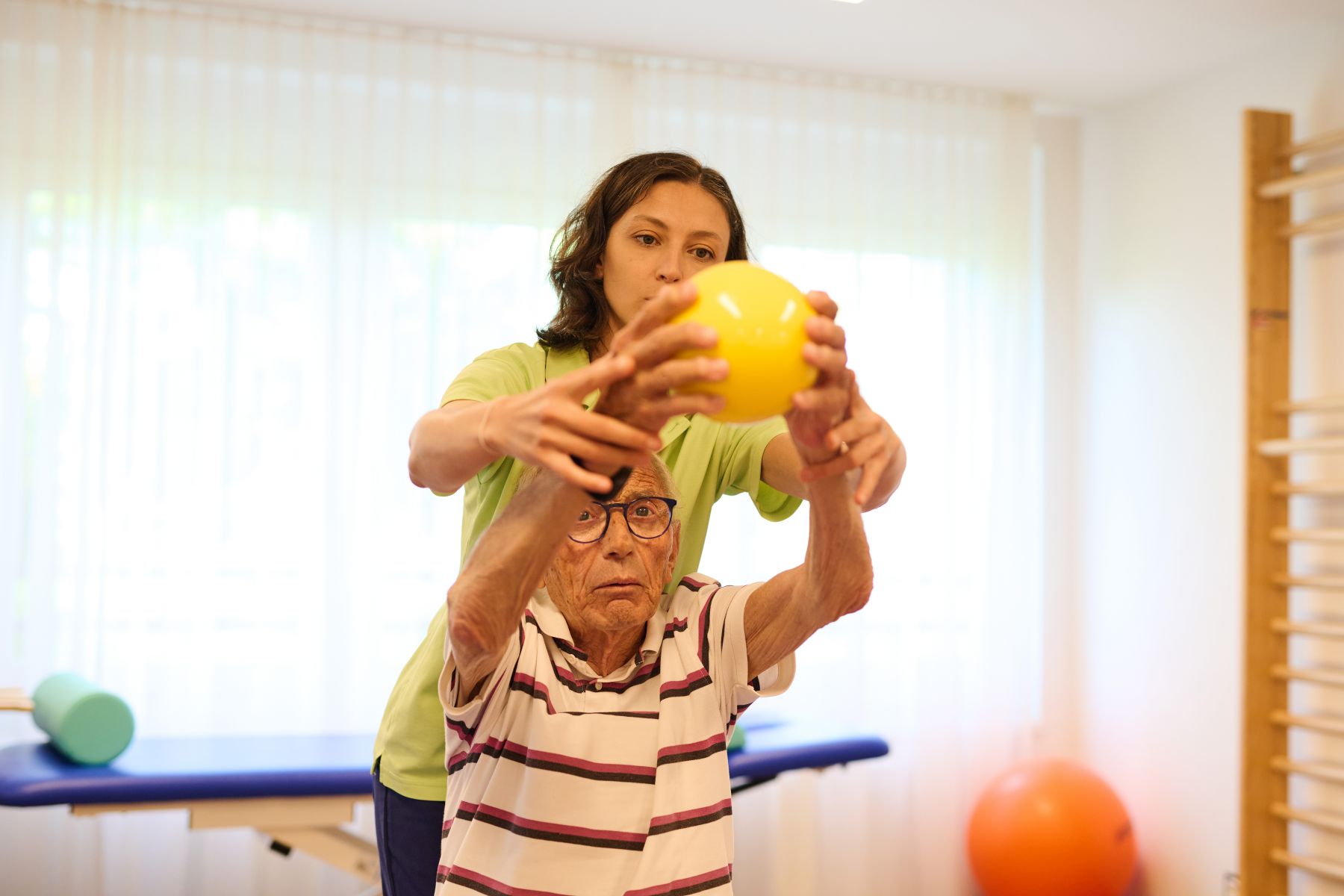 Eine Physiotherapeutin unterstützt einen älteren Herrn, der einen Ball hält.