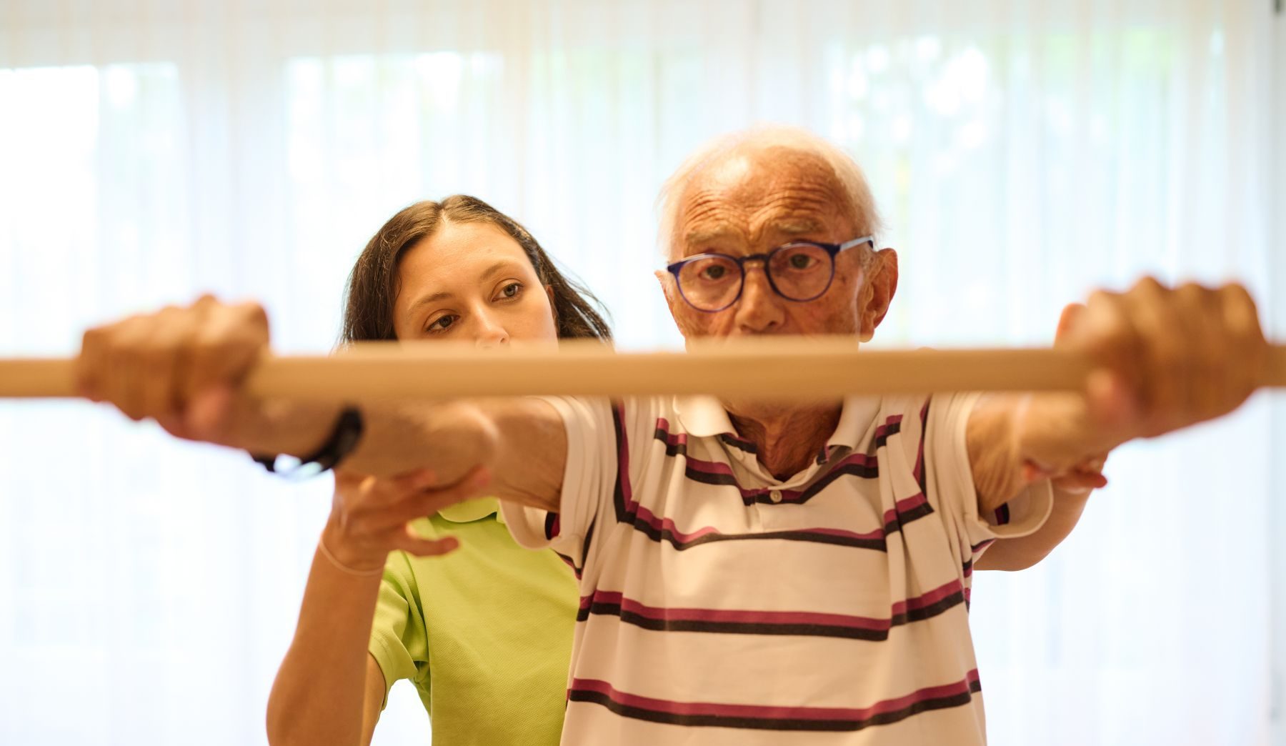 Eine Physiotherapeutin unterstützt einen älteren Herrn, der eine Stange hält.
