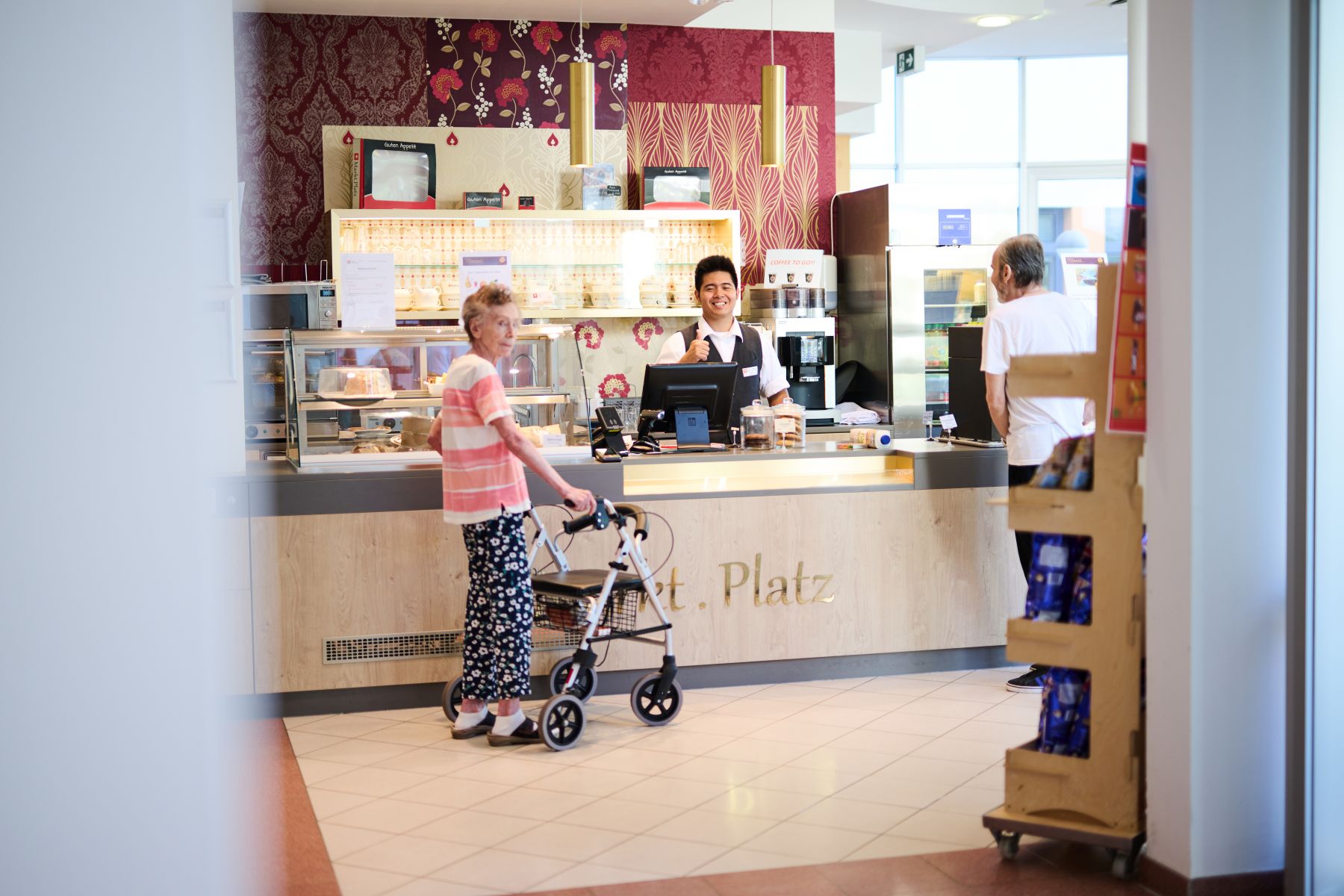 Ein Mitarbeiter, eine Bewohnerin mit Rollator und ein Bewohner am Kiosk "MarktPlatz" im Pensionisten-Wohnhaus Rudolfsheim in Wien.