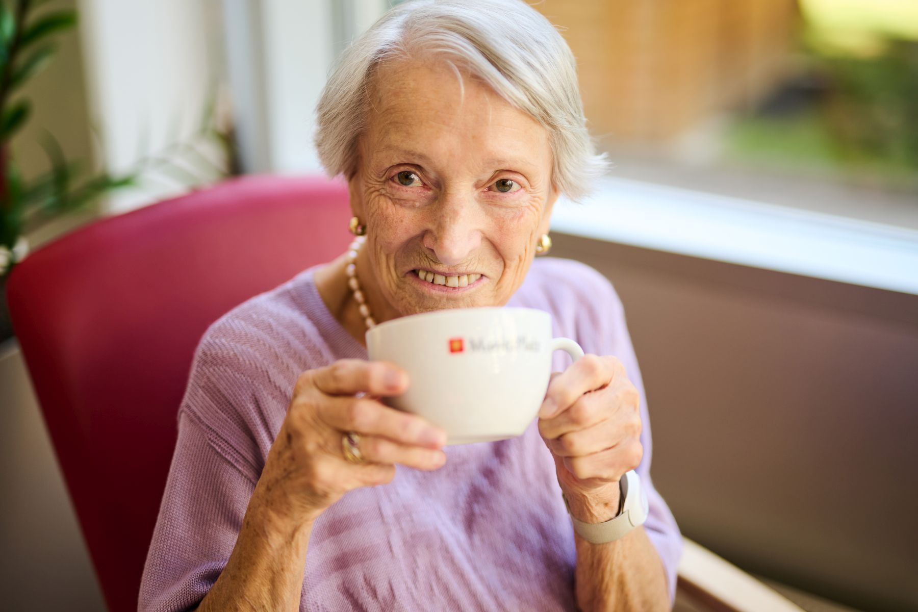 Eine ältere Frau mit kurzen grauen Haaren, Ohrringen und einer Perlenkette trinkt aus einer großen Tasse.
