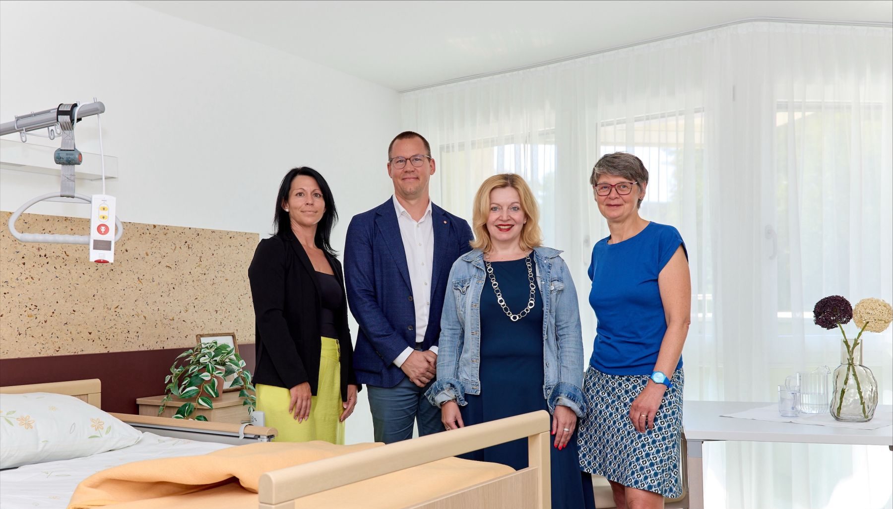 Jasmin Tomassi, Direktorin Haus An der Türkenschanze, Simon Bluma, stellvertretender Geschäftsführer Häuser zum Leben, Anita Bauer, Geschäftsführerin Fonds Soziales Wien (FSW) und Silvia Nossek, Bezirksvorsteherin Wien-Währing stehen hinter einem Bett in einer neuen Pflegestation im Haus An der Türkenschanze.