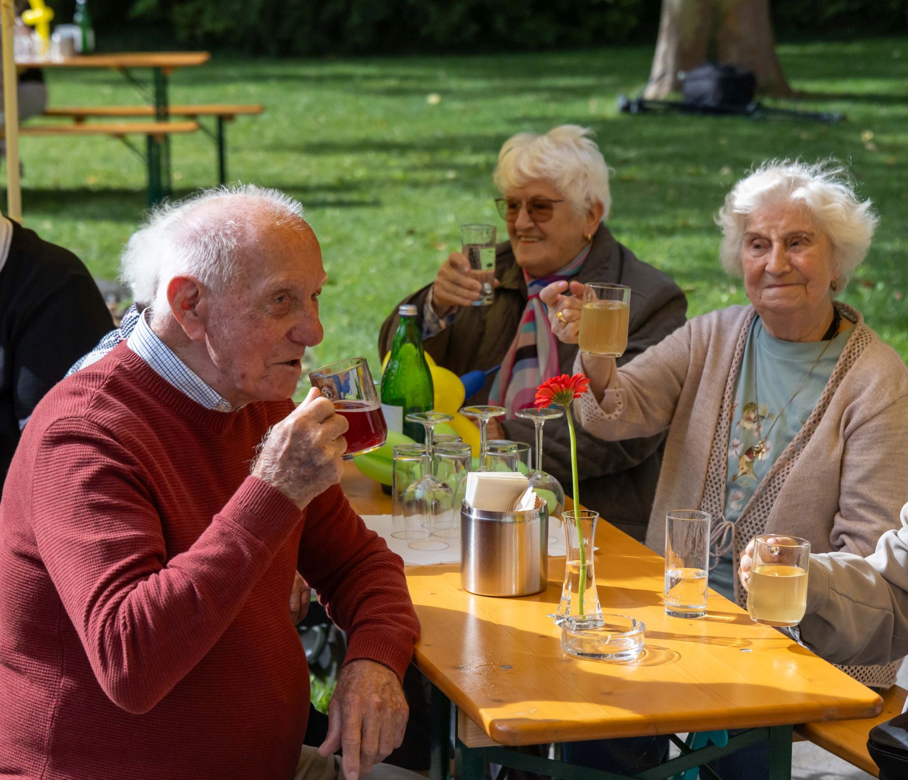 Zwei Seniorinnen und ein Senior sitzen an einem Biertisch im Garten und trinken.