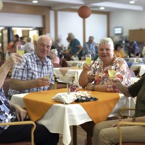 Drei Seniorinnen und ein Senior sitzen an einem Tisch mit orangenem Tischtuch und heben ein Glas.