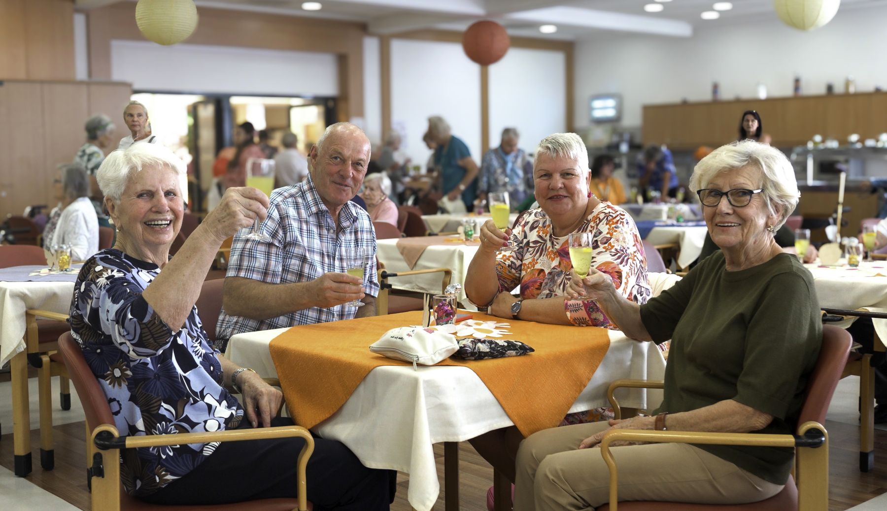 Drei Seniorinnen und ein Senior sitzen an einem Tisch mit orangenem Tischtuch und heben ein Glas.
