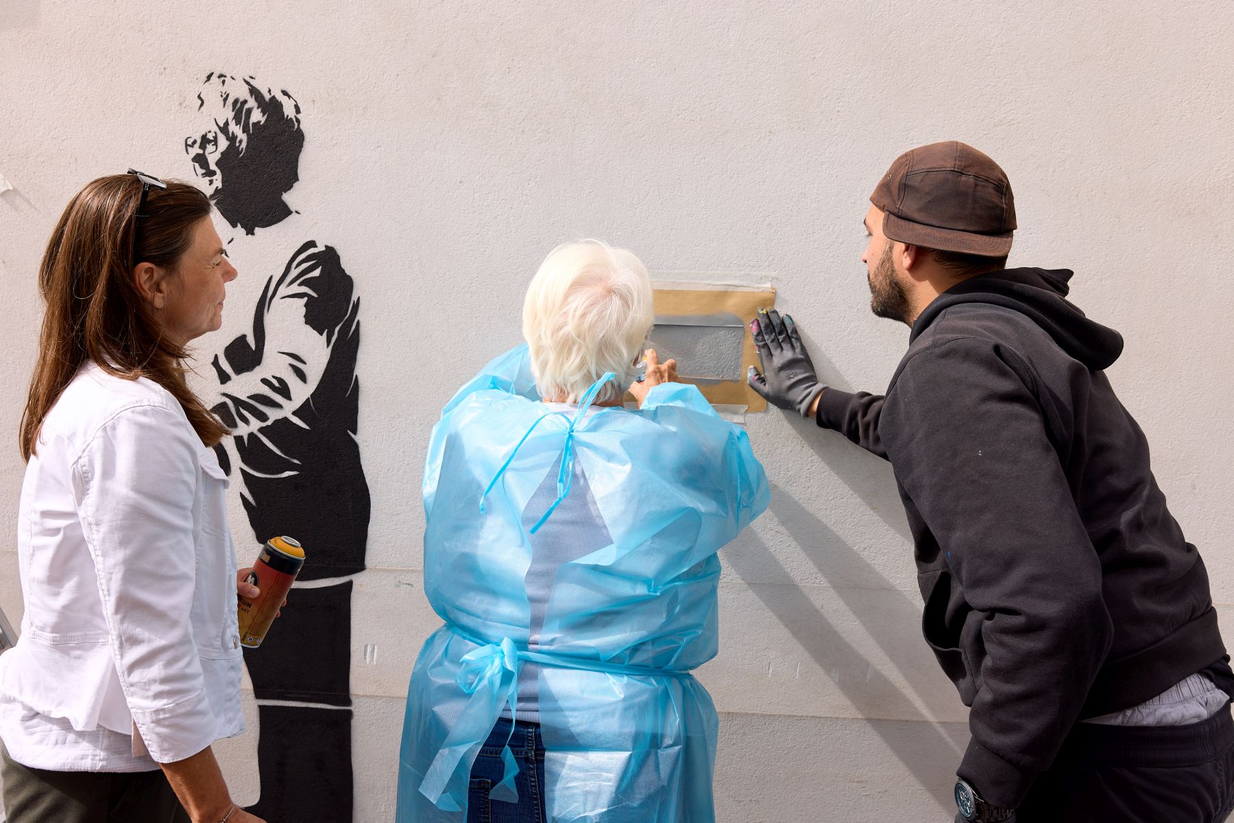 Eine Bewohnerin besprüht mit Hilfe des Graffitikünstlers Robin eine graue Fläche, links Sabine Steinböck (Direktorin Haus Rosenberg)