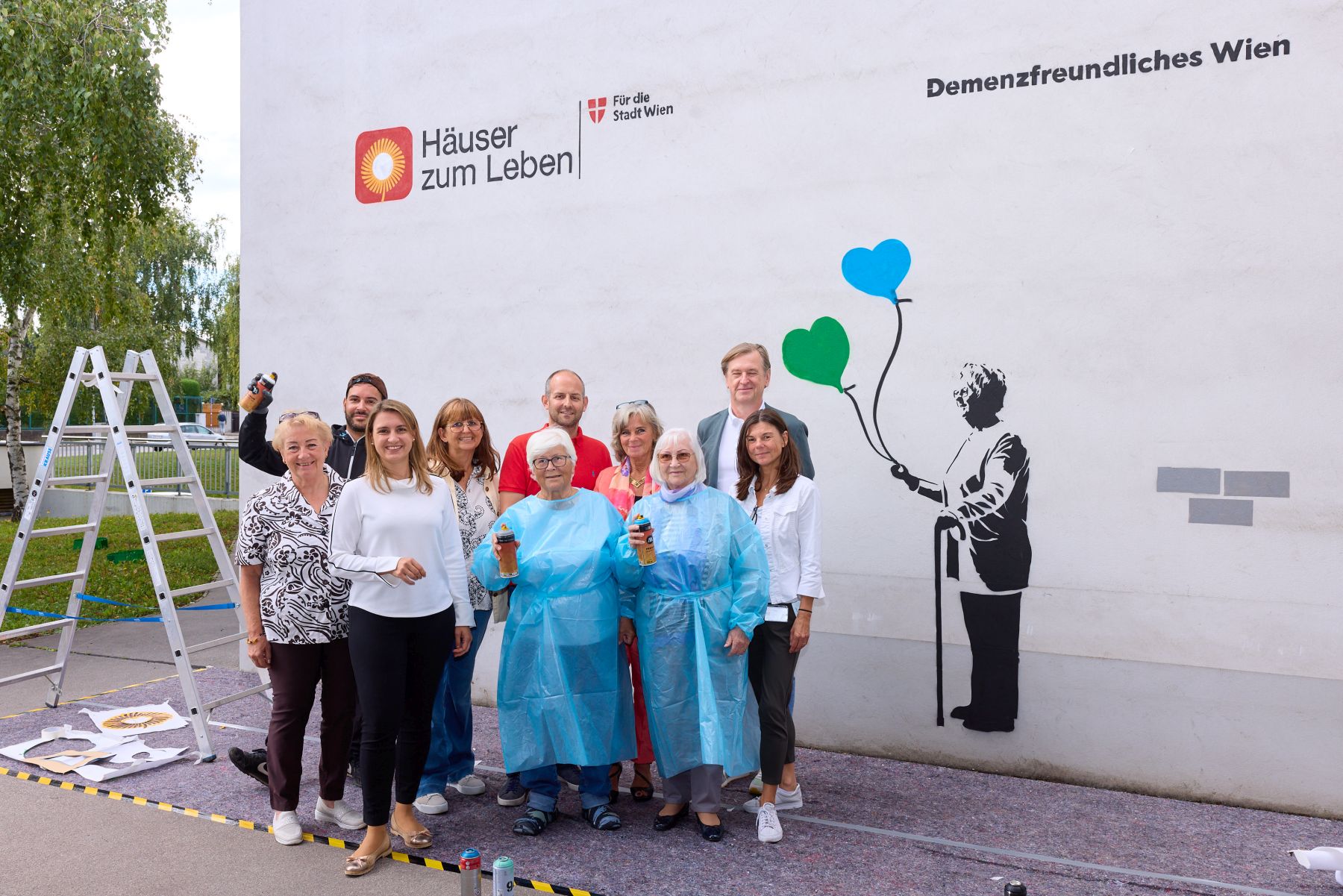 Gruppenbild vor Graffiti mit u.a. Bewohnerinnen Haus Rosenberg, Graffitikünstler Robin Sakar, Evi Pohl-Iser (Häuser zum Leben), Johanna Zinkl (Bezirksvorsteherin Hietzing), Christian Hennefeind (Geschäftsführer Häuser zum Leben), Sabine Steinböck (Direktorin Haus Rosenberg)
