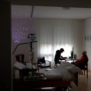 Ein dunkler Raum mit Bett, in dem zwei Männer im Hintergrund an einem Tisch sitzen.