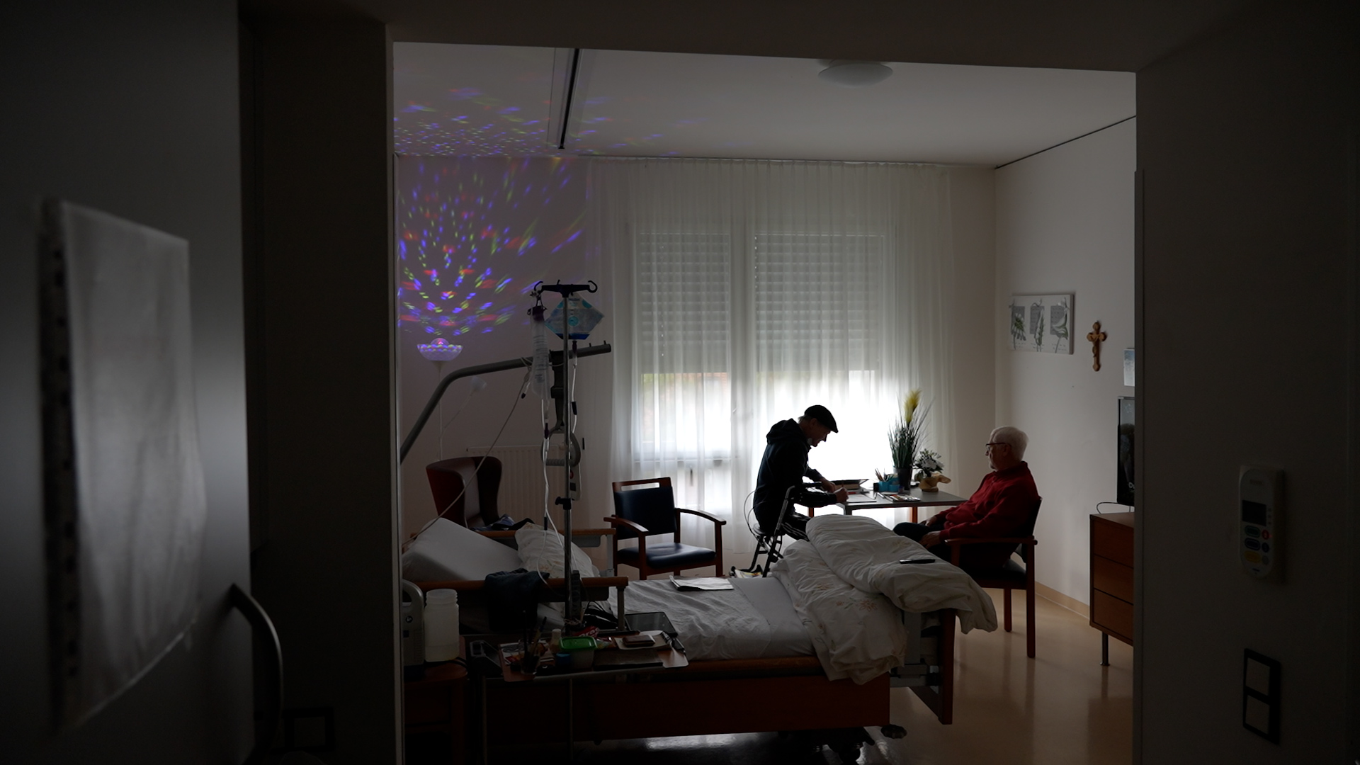 Ein dunkler Raum mit Bett, in dem zwei Männer im Hintergrund an einem Tisch sitzen.