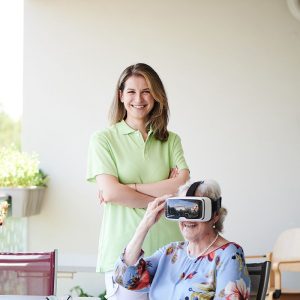 Eine Seniorin mit VR-Brille sitzt an einem Tisch, dahinter steht eine Pflegerin.