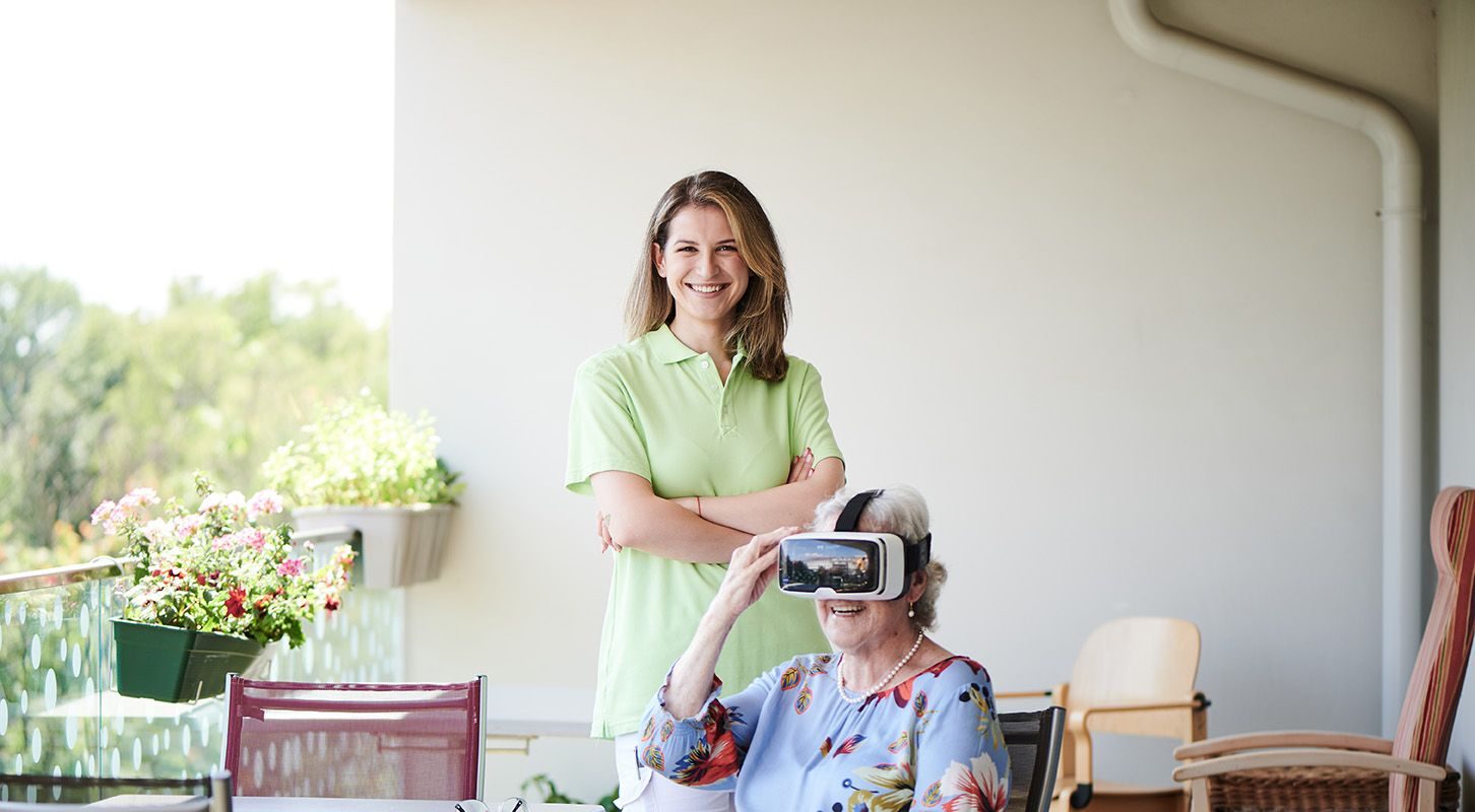 Eine Seniorin mit VR-Brille sitzt an einem Tisch, dahinter steht eine Pflegerin.