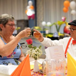 Eine Seniorin und ein Senior prosten sich an einem Tisch zu.