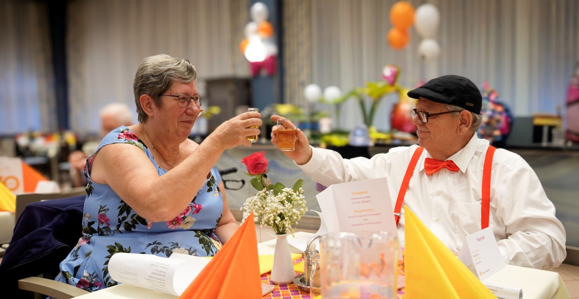 Eine Seniorin und ein Senior prosten sich an einem Tisch zu.