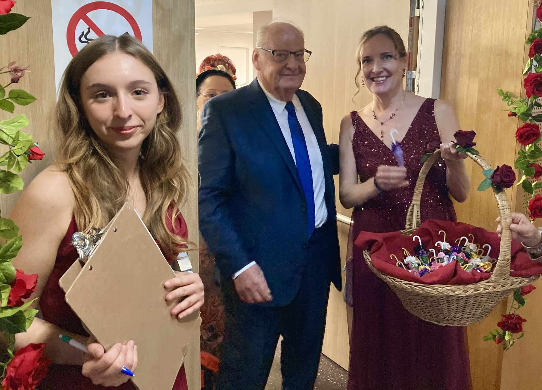 Eine junge Frau am Einlass, ein älterer Herr und Ulrike Geier-Palaschke in rotem Ballkleid und mit Korb beim Winterrosenball im Haus Liebhartstal.
