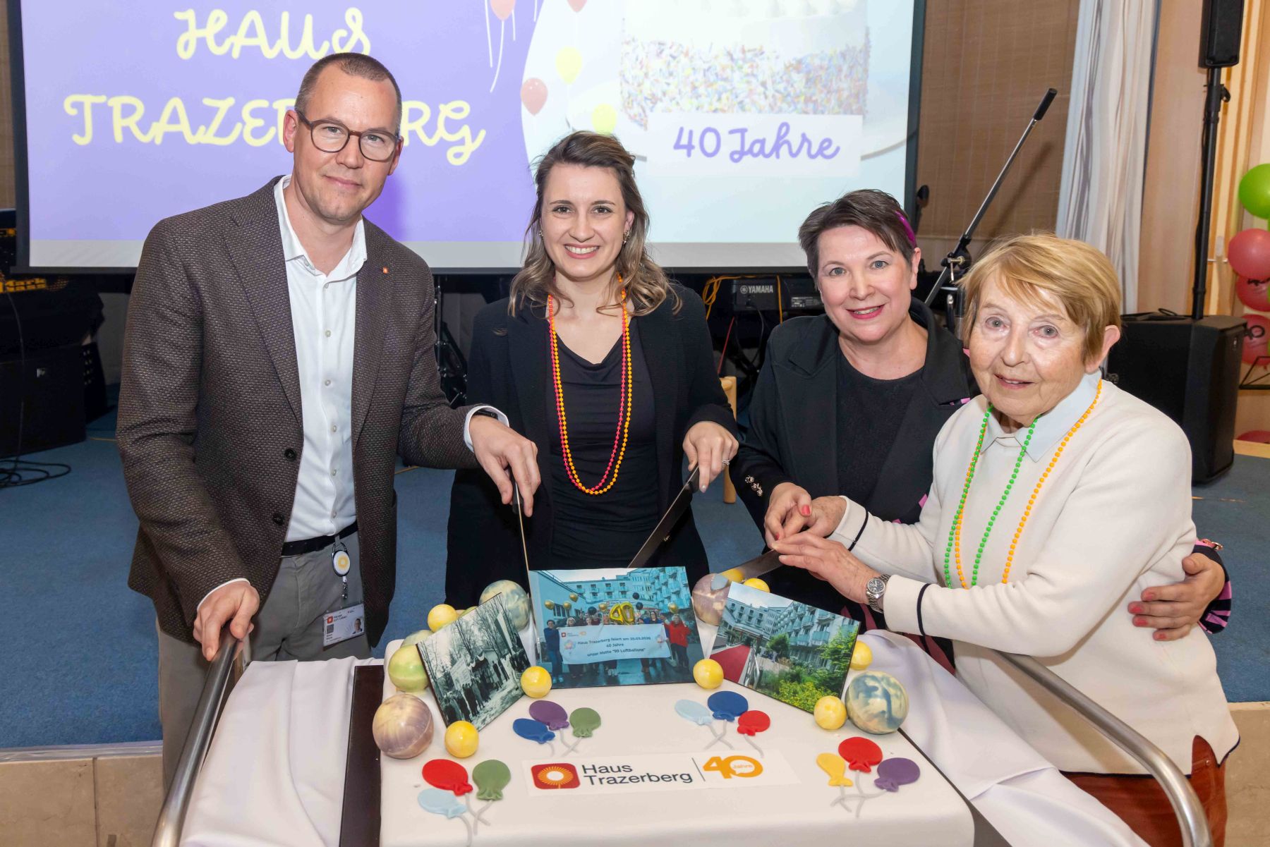 Feier 40 Jahre Trazerberg, Häuser zum Lebern Simon Bluma, stellvertretender Geschäftsführer Häuser zum Leben, Johanna Zinkl. Bezirksvorsteherin Hietzing, Larissa Rebbe, Direktorin Haus Trazerberg und eine Bewohnerin schneiden eine Torte mit Fotos und der Aufschrift "Haus Trazerberg 40 Jahre" an.